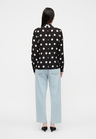 Μαύρη μπλούζα με πουά σχέδιο, μακριά, ελαφρώς φουσκωτά μανίκια, κλείσιμο με κουμπιά στην πλάτη, συνδυασμένη με ανοιχτό μπλε τζιν παντελόνι φαρδύ και μαύρες γόβες.