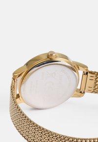 Ρολόι με χρυσό πλέγμα από μπρασελέ, με στρογγυλή κάσα, που διαθέτει υφασμένο πίσω μέρος και κορώνα στην πλευρά. Η επιφάνεια έχει γυαλιστερό φινίρισμα και σκαλιστές λεπτομέρειες.