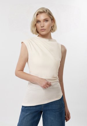 Ξανθιά γυναίκα με κοντά μαλλιά που φοράει μπεζ αμάνικη μπλούζα και μπλε τζιν, στέκεται μπροστά σε ένα απλό ανοιχτό φόντο.