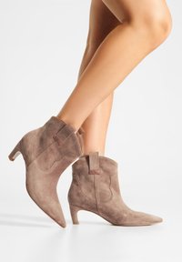 Bottines en daim taupe avec un bout pointu et un talon carré de 5 cm. Équipées d'une languette et de détails de couture subtils de style western.