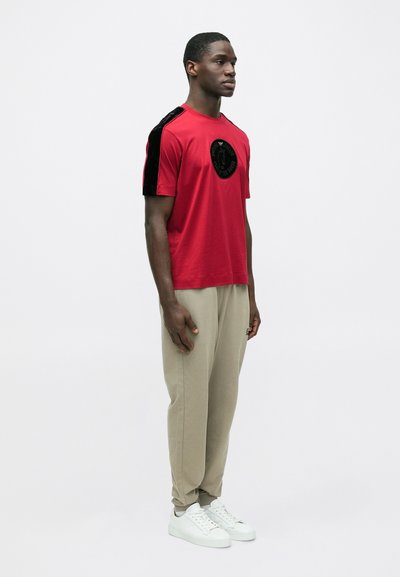 Rdeča majica s kratkimi rokavi s črnimi žametnimi poudarki na ramenih in okroglim logotipom; združena z bež hlačami in belimi supergami.