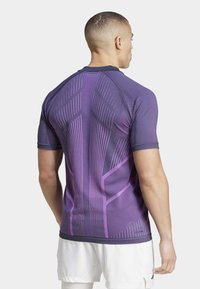 Camiseta deportiva púrpura con líneas y patrones gráficos, de manga corta y un diseño ajustado. El material parece suave y elástico.