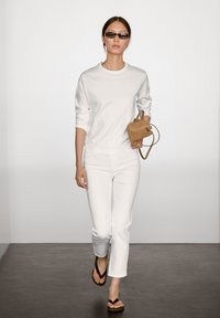 Bílá košile s dlouhým rukávem, bílé džíny, černé žabky, nese malou béžovou semišovou kabelku. Modelka kráčí na neutrálním pozadí.