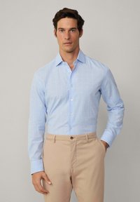 Camisa abotonada de color azul claro con un estampado a cuadros, que presenta mangas largas y un cuello clásico. Combinada con pantalones beige.