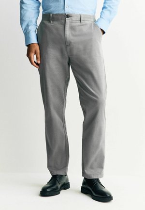 Man wearing light blue long-sleeve shirt, gray dress pants, and black leather shoes, standing with one hand in pocket.