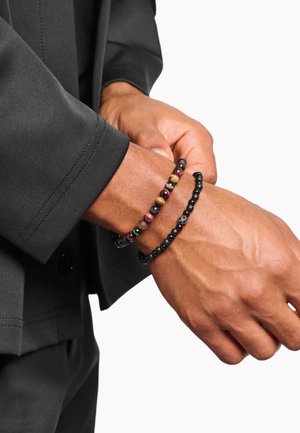 Due braccialetti su un polso: uno con perline rotonde multicolori, l'altro con piccole perline nere e un piccolo ciondolo d'argento, indossato con una giacca nera.