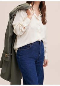 Blouse beige boutonnée avec manches longues et poignets, associée à un jean en denim bleu. Un trench-coat vert est posé sur l'épaule.