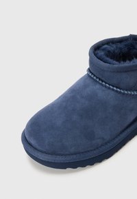 Botas de tobillo de ante azul con un interior suave similar al de la piel de un animal, punta redondeada y suela texturizada. Presenta detalles de costura alrededor de la parte superior.
