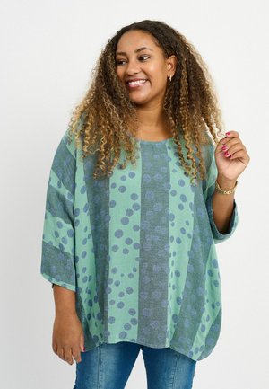 Blusa verde de gran tamaño con lunares azules y rayas, con mangas tres cuartos y cuello redondo; tejido suave y ligero.