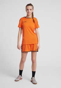 Orange sports trøje og shorts med sorte accenter. Trøjen har korte ærmer og rund hals; shortsene har elastisk linning.