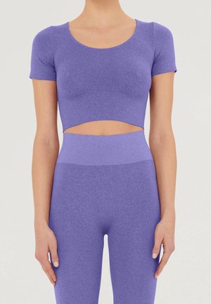 Femme portant un haut court lavande à manches courtes et un legging taille haute assorti sur un fond clair uni.