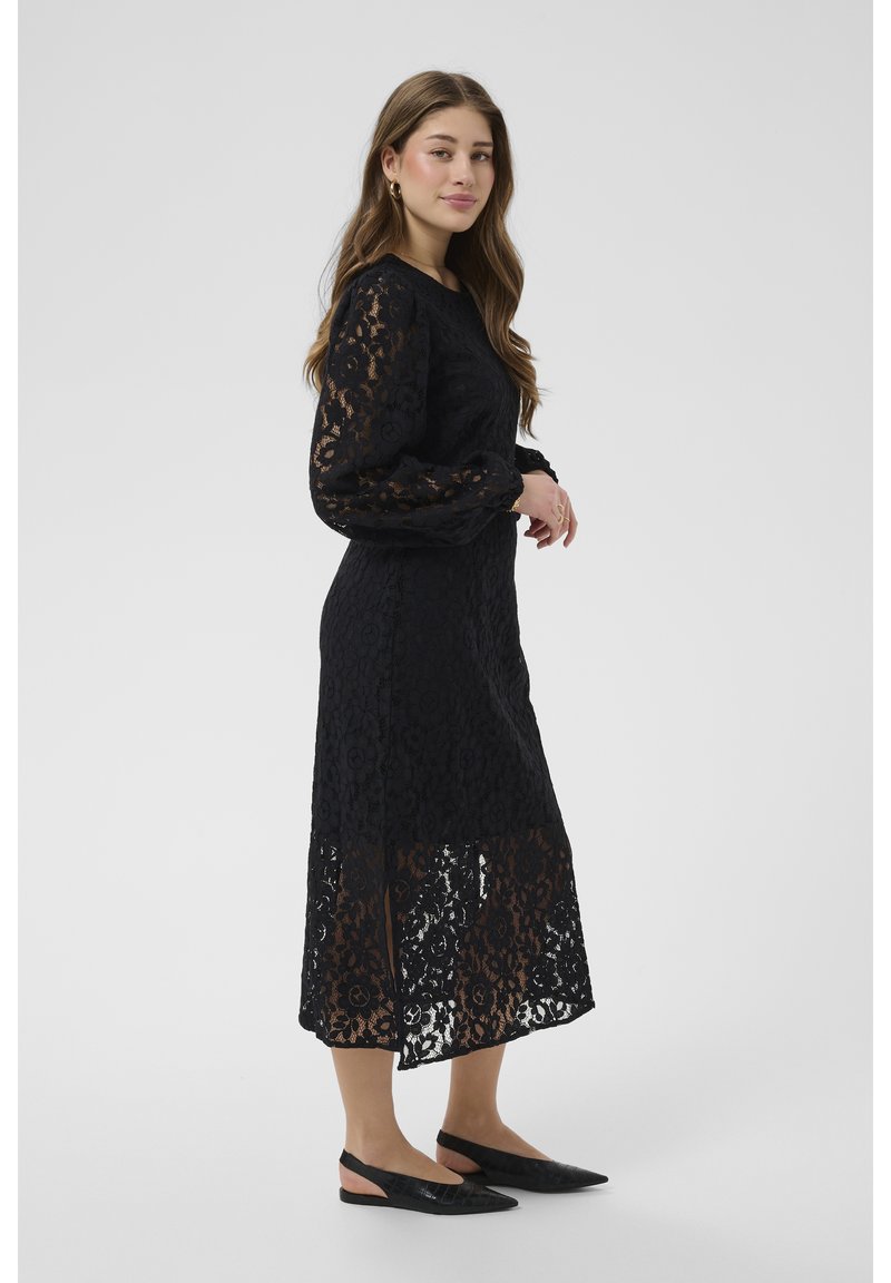 Jauna sieviete, redzama no sāniem, valkājot garu melnu mežģīņu kleitu ar garām piedurknēm un melnas, stikla papēžu aizmuguri atvērtas kurpes ar smailiem purngaliem.