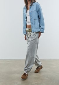 Personne portant une veste en denim bleu clair, un crop top blanc, une ceinture en foulard à motifs, un pantalon de survêtement gris et des chaussures décontractées marron marchant à l'intérieur.