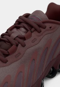 Burgundowe buty sportowe z teksturowanym wykończeniem, siateczkowymi akcentami i charakterystycznym wzorem wycięć po bokach. Widoczne sznurówki i logo.