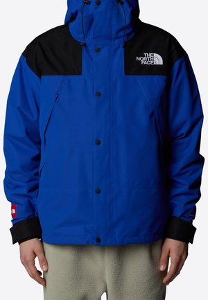 Niebiesko-czarna kurtka North Face z guzikami z przodu, regulowanym kapturem i logo na lewej piersi, noszona z jasnoszarymi spodniami z polaru.