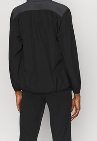 Zwarte windbreakerjas met een grijze schouderpas, elastische manchetten en tailleband. Gladde, lichte stof met een functioneel ontwerp.