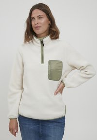 Fleeceový pulóver v odtieňoch off-white, so zeleným zipsovým vreckom a golierom. Má voľný strih a rebrované manžety. Mäkkučká textúra po celom obvode.