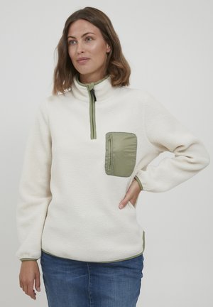 Fleece pullover in off-white, with a green zippered pocket and collar. Features a relaxed fit and ribbed cuffs. Soft texture throughout.