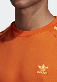 Camiseta de algodón naranja con cuello redondo, que presenta tres rayas horizontales en los hombros y un pequeño logo blanco de Adidas en el pecho.