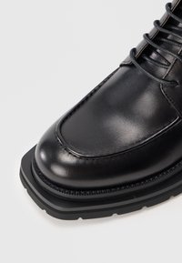 Zapato negro de piel con cordones, punta cuadrada y suela de goma gruesa sobre un fondo gris.