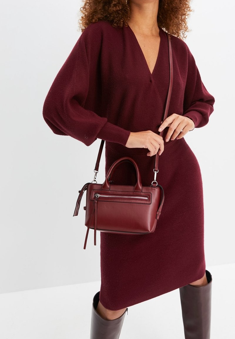 Femme portant une robe en maille bordeaux avec des manches ballon, un sac à main en cuir bordeaux assorti, et des bottes marron montant jusqu'aux genoux.