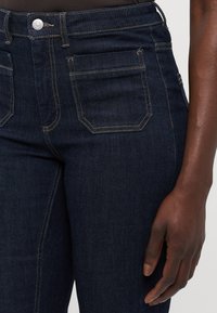 Tamsios džinsinės kelnės turi prigludusį modelį su aukštu liemeniu, matomu siuvinėjimu ir kontrastingomis aukso detalėmis. Klasikinis penkių kišenių stilius.