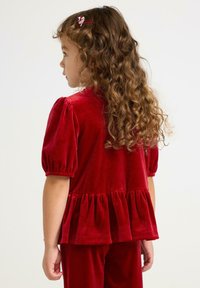 Jong kind met lang krullend haar, gekleed in een rode fluwelen jurk met pofmouwen en een gerimpelde zoom, staande in profiel tegen een effen achtergrond.