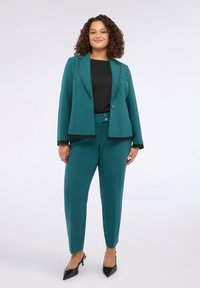 Blazer teal con bottoni neri decorativi, abbinato a pantaloni a gamba affusolata dello stesso colore e una camicia nera. L'outfit presenta una texture di tessuto liscia.