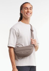 Sac en bandoulière matelassé marron avec sangle réglable porté en travers du corps, featuring un logo discret. Texture lisse et design curvé et minimaliste.
