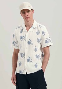 Camicia a manica corta con bottoni in lino bianco con motivo floreale blu. Presenta un colletto a campana e bottoni semplici. Indossata con pantaloni scuri.
