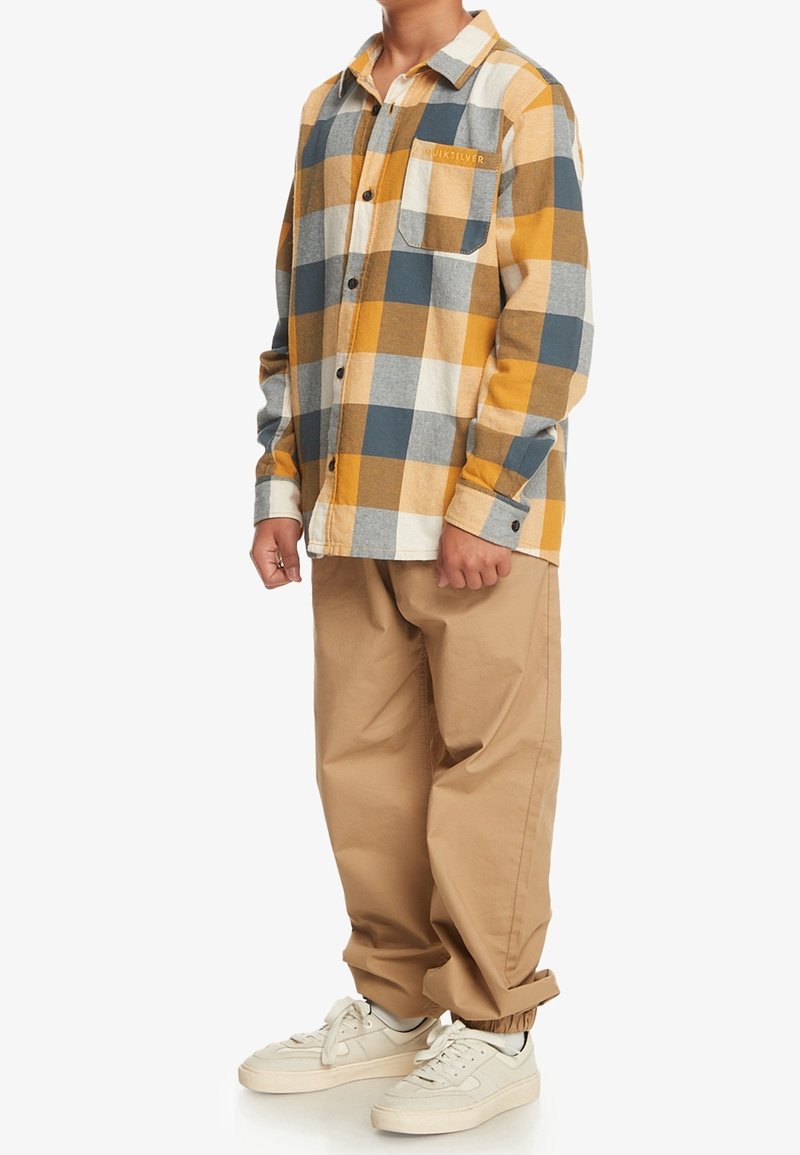 Personne portant une chemise à carreaux mustard, grise, blanche et marron avec une poche poitrine, un pantalon beige et des baskets blanches, debout sur un fond blanc.