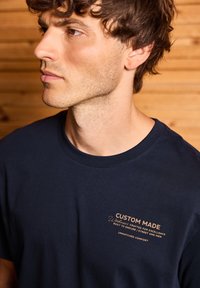 Mörkbl�å bomullströja med korta ärmar och rundad hals, med guldtext på vänster bröstområde som säger "CUSTOM MADE."