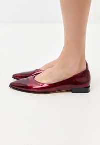 Chaussures plates en cuir verni rouge avec un bout pointu, un petit talon noir et un design découpé sur les côtés. Texture lisse et brillante.