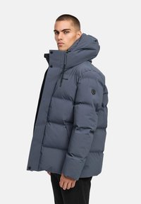 Det vadderade grå jackan har en hög krage, justerbar huva och en logotypapplikation. Tyget har en slät yta med synliga sömmar.