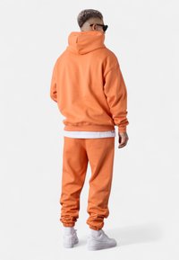 Oranger Hoodie mit Kängurutasche, passende Jogginghosen mit gerippten Säumen, getragen mit weißen Sportschuhen. Glattes Material, lockere Passform.