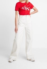 Červené tričko s krátkým rukávem a logem Calvin Klein, kombinované s bílými vysokými kalhotami širokého střihu a bílými teniskami. Jednoduché pozadí.