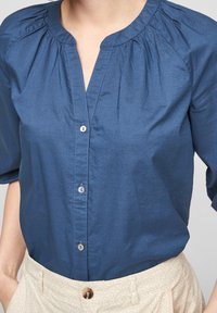 Mujer con una camisa de botones azul con tres botones visibles y pantalones color crema con un sutil patrón geométrico.