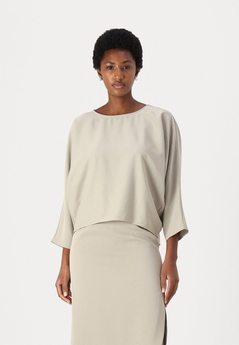 Top grigio chiaro, dalla vestibilità morbida e con maniche lunghe, con un ampio scollo, indossato sopra una gonna coordinata. Tessuto morbido con una silhouette leggermente drappeggiata.