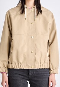 Beżowa, wodoodporna kurtka z kapturem, z zatrzaskami, elastycznymi mankietami, dwoma przednimi kieszeniami oraz ściągaczem u dołu. Gładka faktura.