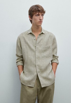 Light beige button-up shirt with a textured fabric, rolled sleeves, and a chest pocket. Worn with matching beige trousers.