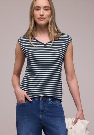 Ung kvinde med langt blondt hår iført en navyblå og hvidstribet ærmeløs top og blå jeans, der holder en let jakke i den ene hånd.