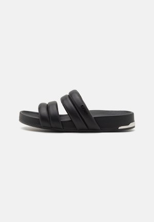Sort slidesandal med tre polstrede remme og flad sål med en lille sølvdetalje ved hælen, vist fra siden mod en ensfarvet baggrund.