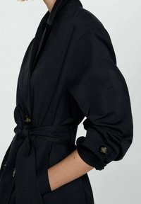 Trench-coat noir en tissu doux. Doté d'une ceinture à la taille et de poignets retroussables avec des boutons distinctifs. Texture lisse et coupe ajustée.