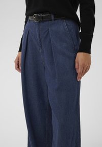 Közeli kép egy személyről, aki magas derekú, sötétkék farmernadrágot visel redőkkel és fekete bőrövvel, fekete hosszú ujjú felsővel párosítva.