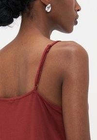 Camisole rusti punarus dengan tali tipis, kain ringan, dan tekstur halus. Memiliki leher bulat dan tali yang dapat disesuaikan.