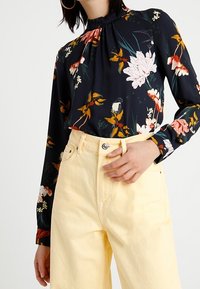 Blusa nera a fiori con collo alto, maniche lunghe e design arricciato, abbinata a pantaloni gialli a vita alta.