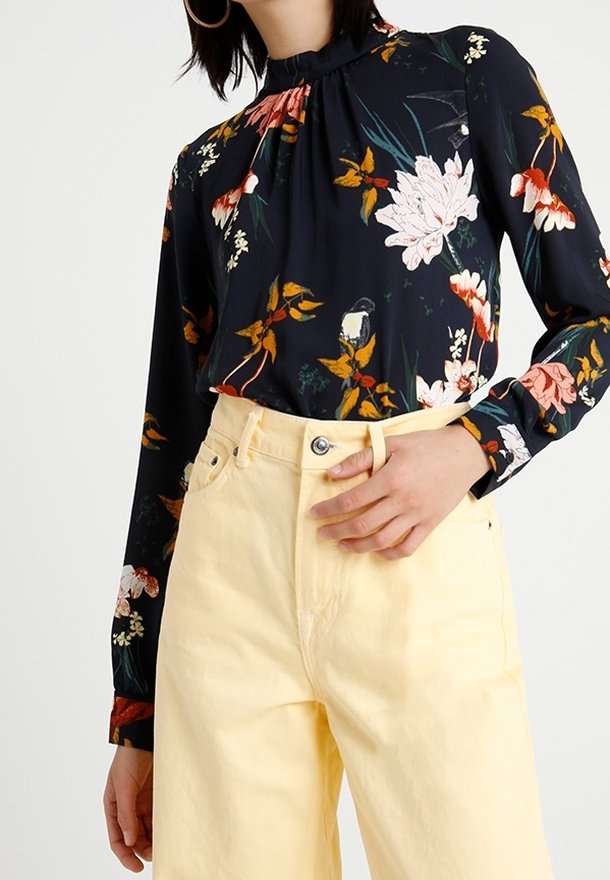 Blusa nera a fiori con collo alto, maniche lunghe e design arricciato, abbinata a pantaloni gialli a vita alta.