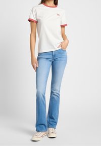 T-shirt blanc avec des bordures rouges, logo sur la poitrine, associé à un jean léger évasé bleu clair et des sneakers beiges.