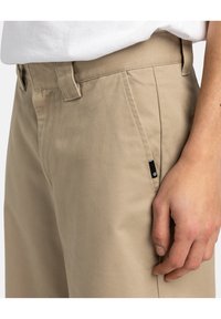 Pantaloni di cotone beige con una trama liscia, dotati di tasca laterale, passanti per la cintura e un piccolo etichetta nera vicino alla tasca.