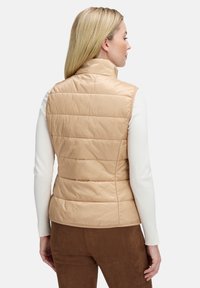 Beige puffer vest met hoge kraag, voorzien van verticale stiksels en een gladde, glanzende textuur, gedragen over een wit langarmshirt.